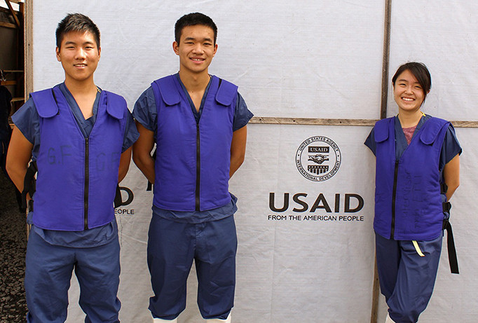 Three young adults smile and pose in front of a wall imprinted with the USAID logo