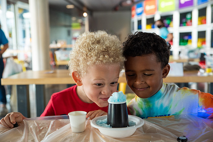 Two young kids stare eagerly at cup filled with blue crushed ice inside a bright and colorful workshop