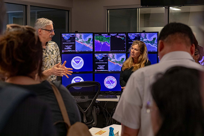 Man presents to small group of visitors with nine monitors showing earthquake detections behind him