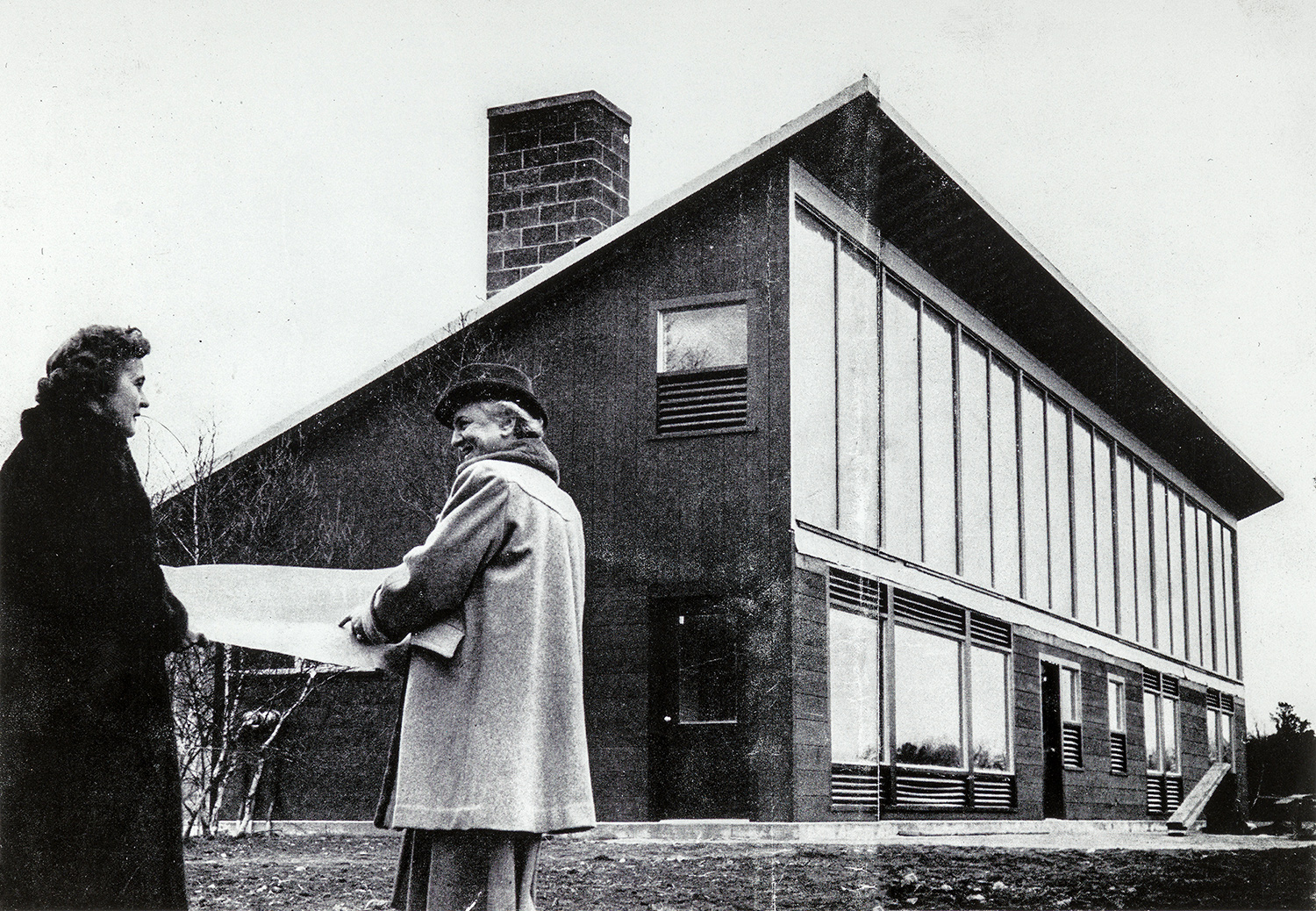 Maria Telkes and Eleanor Raymond in front of the Dover Sun House, holding a blueprint for the house between them. Raymond points to something in it as she smiles at Telkes.