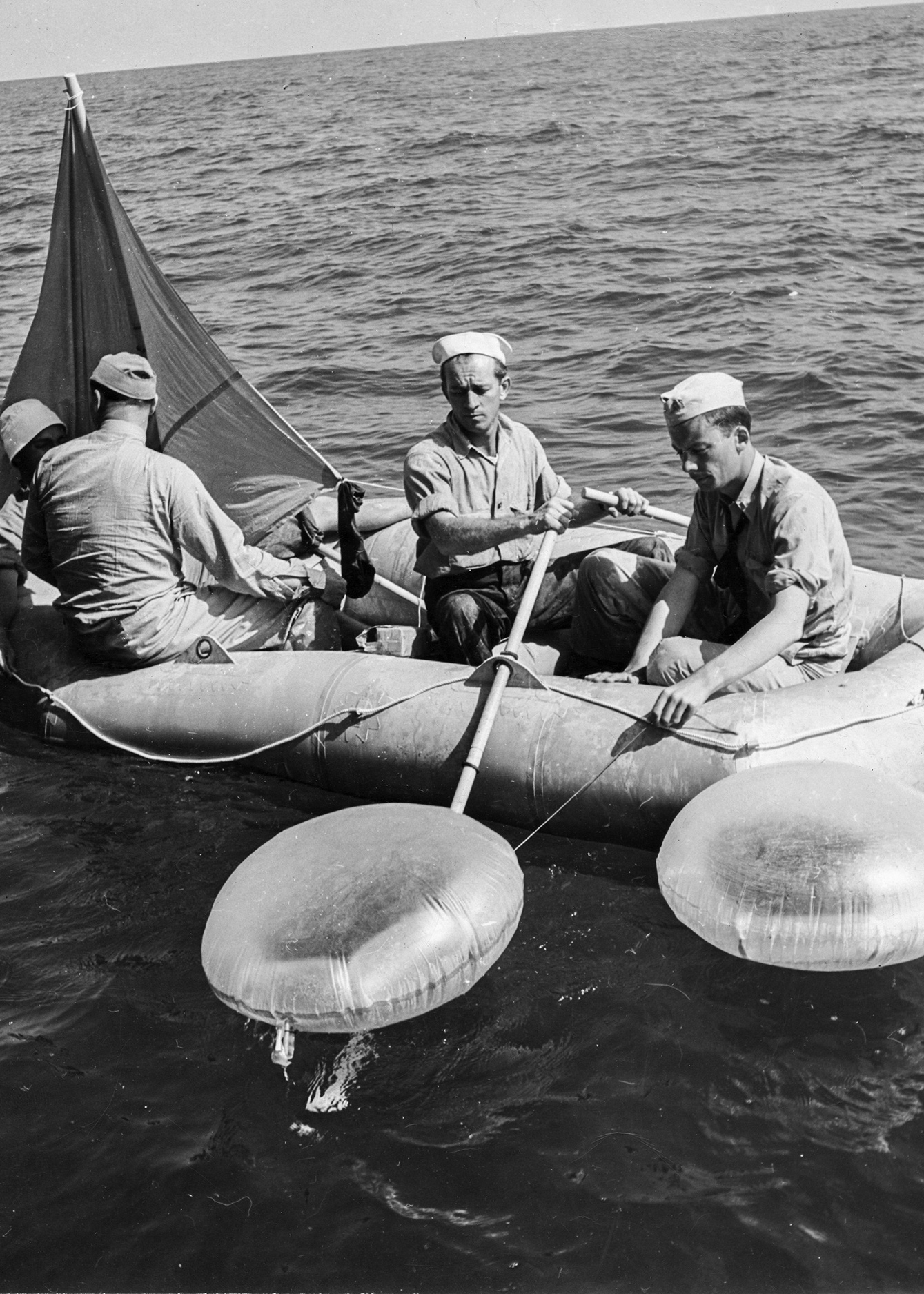 In a black-and-white, mid-20th century image, three uniformed men on a life raft look at two balloon-like containers floating on the water close to their raft.