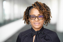 Percina, a woman with light brown skin, short brown-black curly hair, glasses, and dangling silver earrings smiles in a sunny USPTO headquarters hallway.