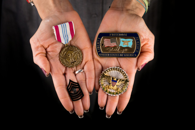 hands holding a Defense Meritorious Service Medal, a Challenge Coin from Secretary of Defense Donald Rumsfeld, an Office of the Secretary of Defense Identification Badge, and Master Gunnery Sergeant rank insignia.