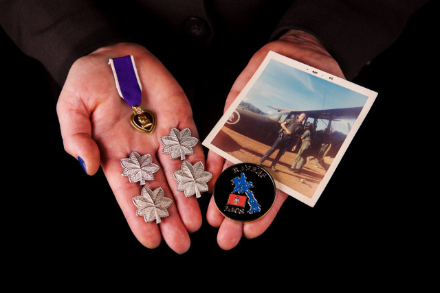 hands holding Purple Heart, a November 1965 photo of soldier loading radios into an O1E aircraft in Laos during the Vietnam War, a Ravens coin presented to soldier at a Ravens annual reunion, and out lieutenant colonel rank insignia