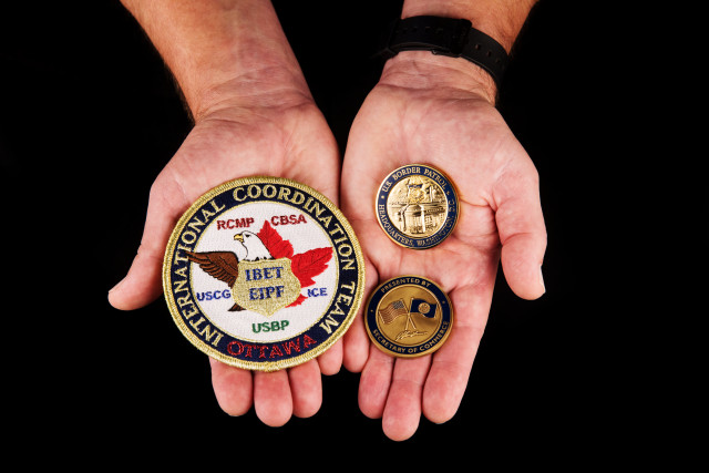 hands holding Challenge coins from the Secretary of Commerce and Chief, U.S. Border Patrol, and a patch from the United States, Canada Integrated Border Enforcement Team (IBET). 