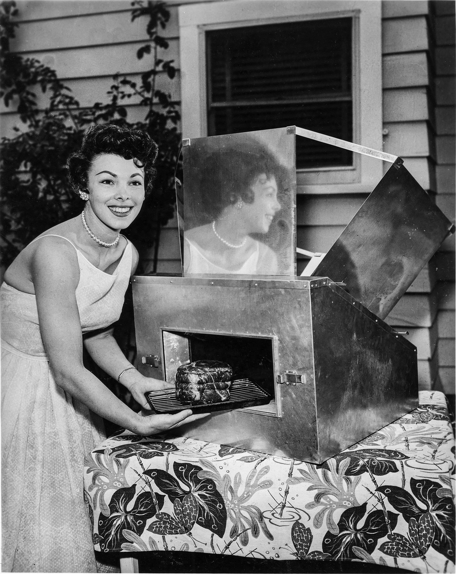 A black-and-white photo shows a mid-20th century woman smiling at the camera as she pulls a meal out a solar oven.