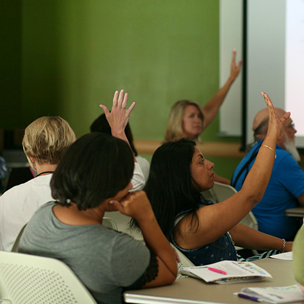 A group of teachers seated in a classroom raising their hands to ask questions
