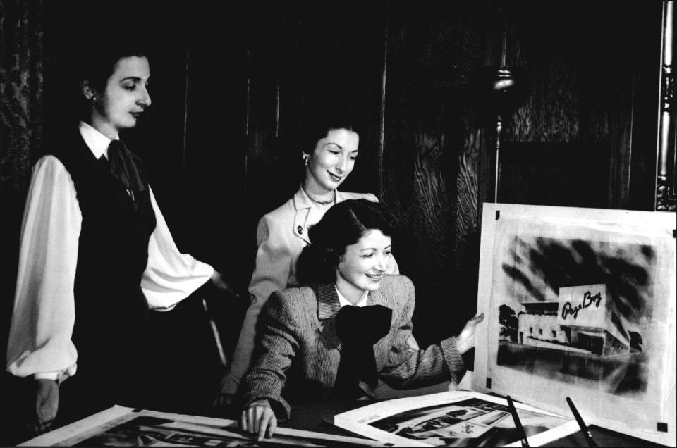 Three fashionably dressed white women surrounding a desk, posing and admiring posters of Page Boy store fronts