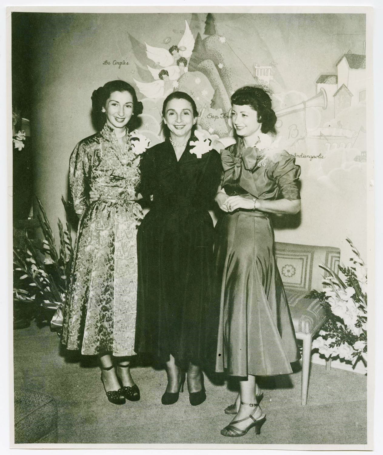 Three white women standing in long-sleeve, ankle-length cocktail dresses, with linked arms, posing and smiling