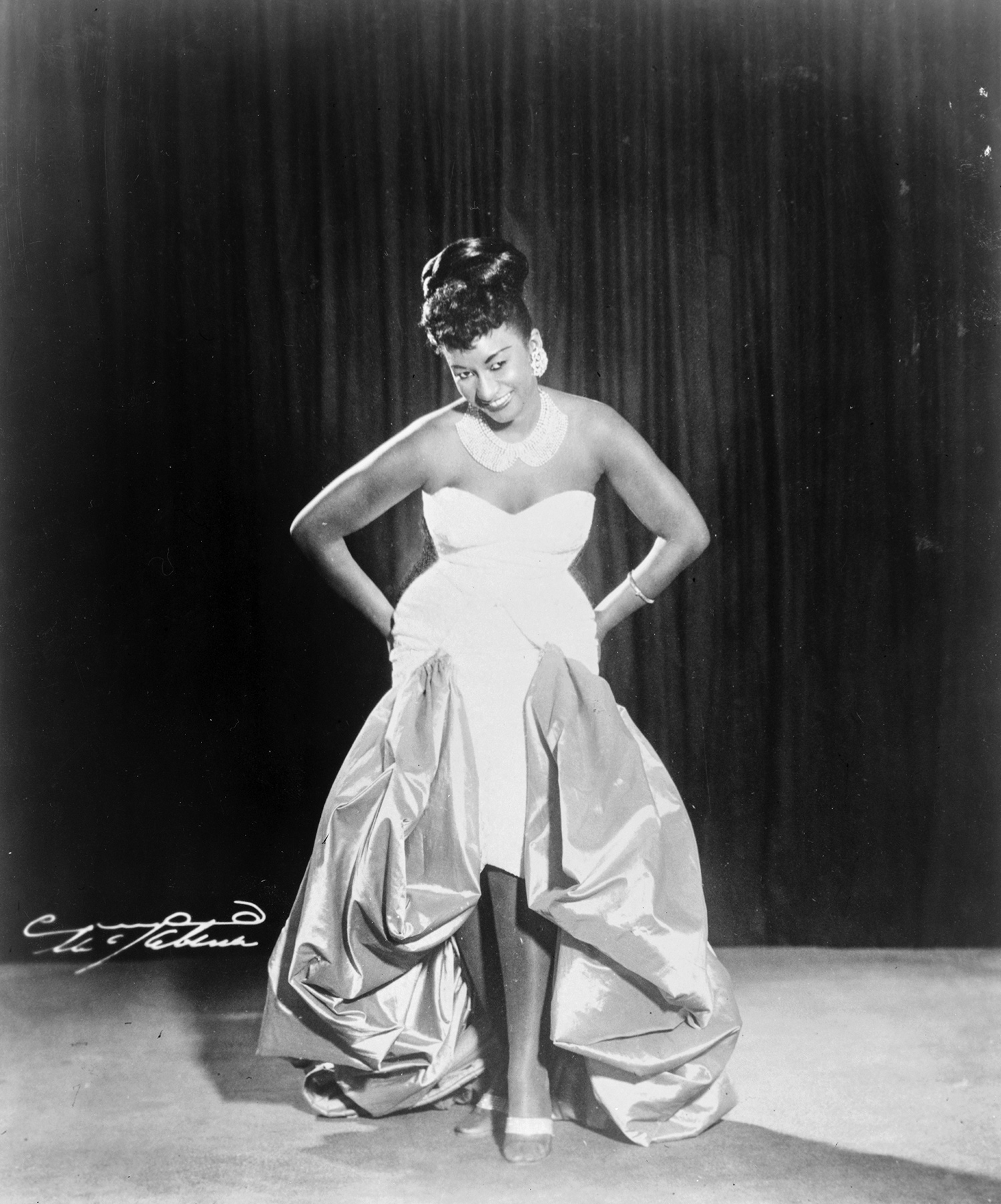 A full-length photographic portrait of a woman in a glamorous evening dress and smiling, her gaze cast downward. 