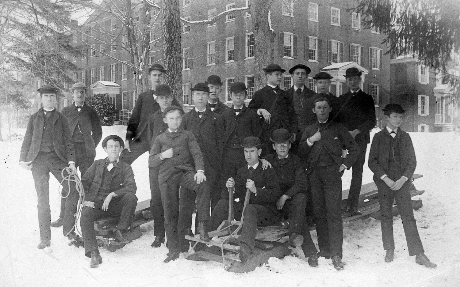 Group portrait of teenaged boys, some on large sleds, standing in front of a brick school building. 