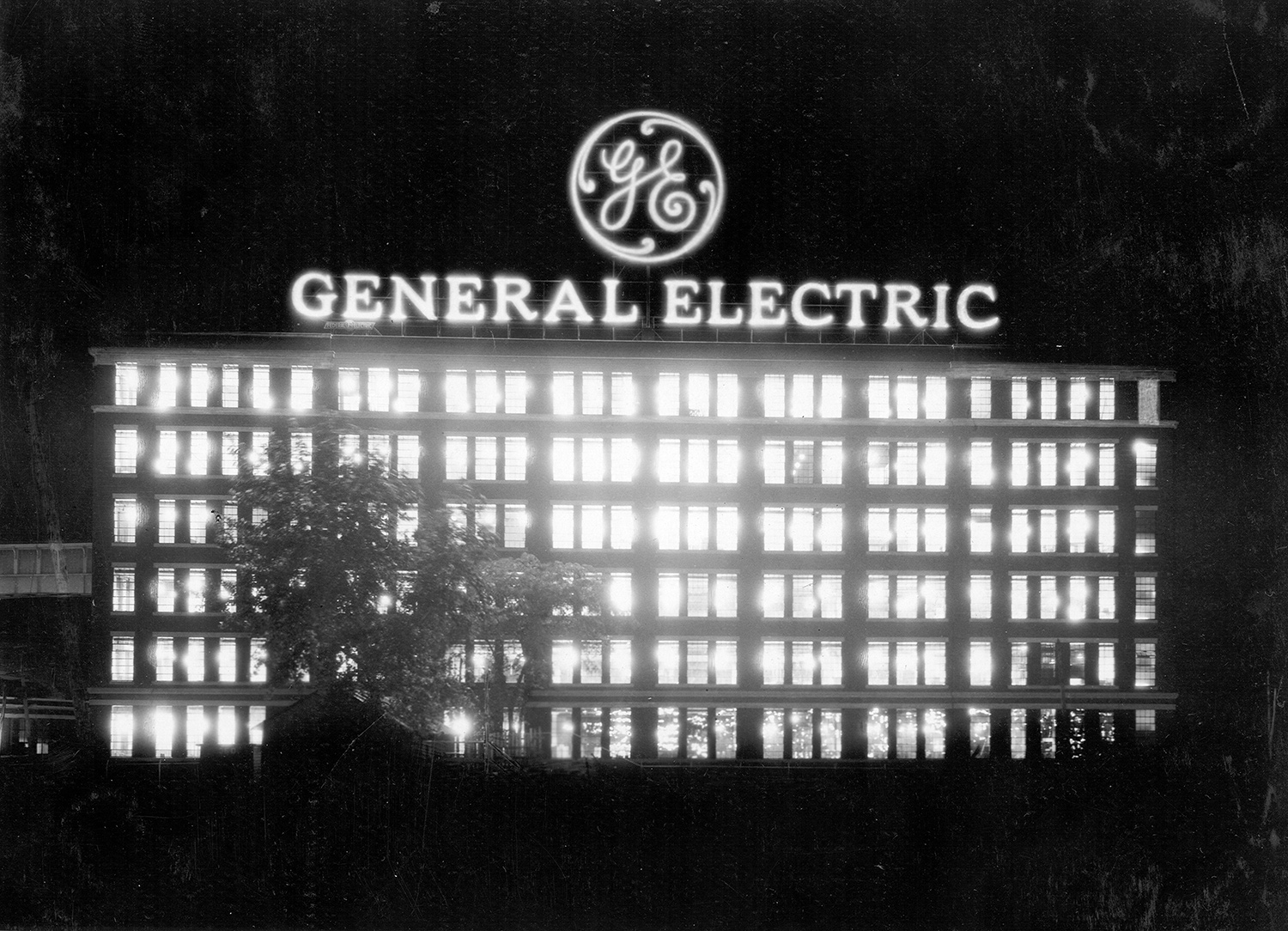 A large commercial building at night, with hundreds of windows, all illuminated. An illuminated logo and sign at the top of the building reads, “GE: General Electric.” 