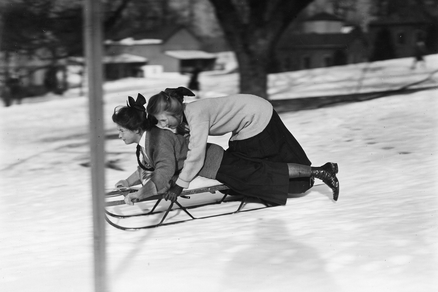 Two girls wearing long skirts and bows, on a sled, going downhill. 