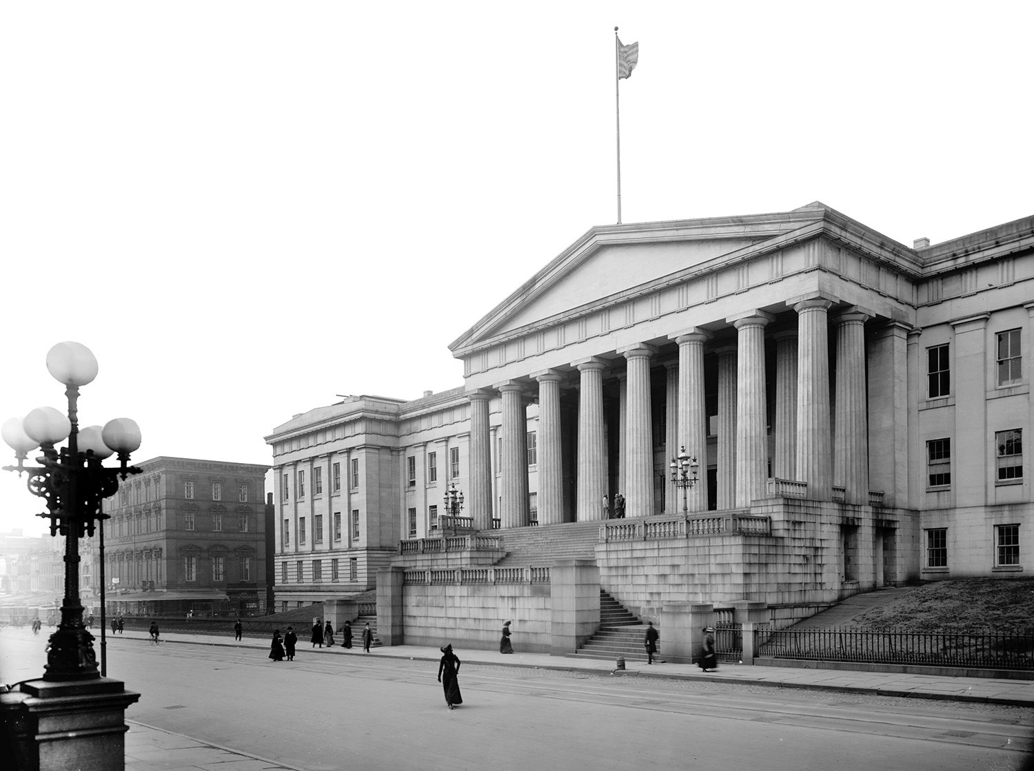 Black and white photo of a large government building with columns in the front and an American flag waving on the roof