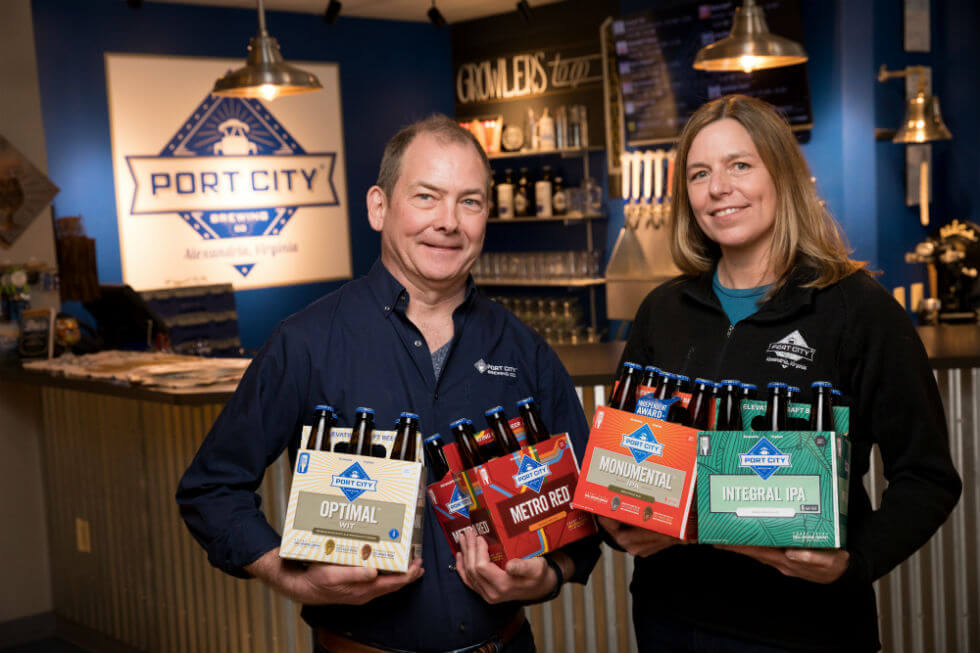 Image: Port City® Brewing Company founders Bill and Karen Butcher in the brewery’s taproom hold six packs of some of their registered trademark beers, including, from left, Optimal® Wit, Metro Red®, Monumental® IPA, and Integral® IPA.
