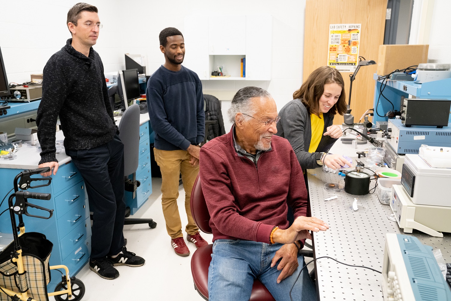 Jim West seated in his lab and surrounded by three students