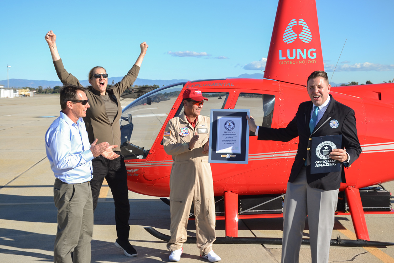 Martine Rothblatt(excitedly jumping) and the team from Tier one engineering proudly celebrating in front red Electric helicopter that, with the word lung written on it, huge smile, that just set Guinesss World Record being awarded plaque