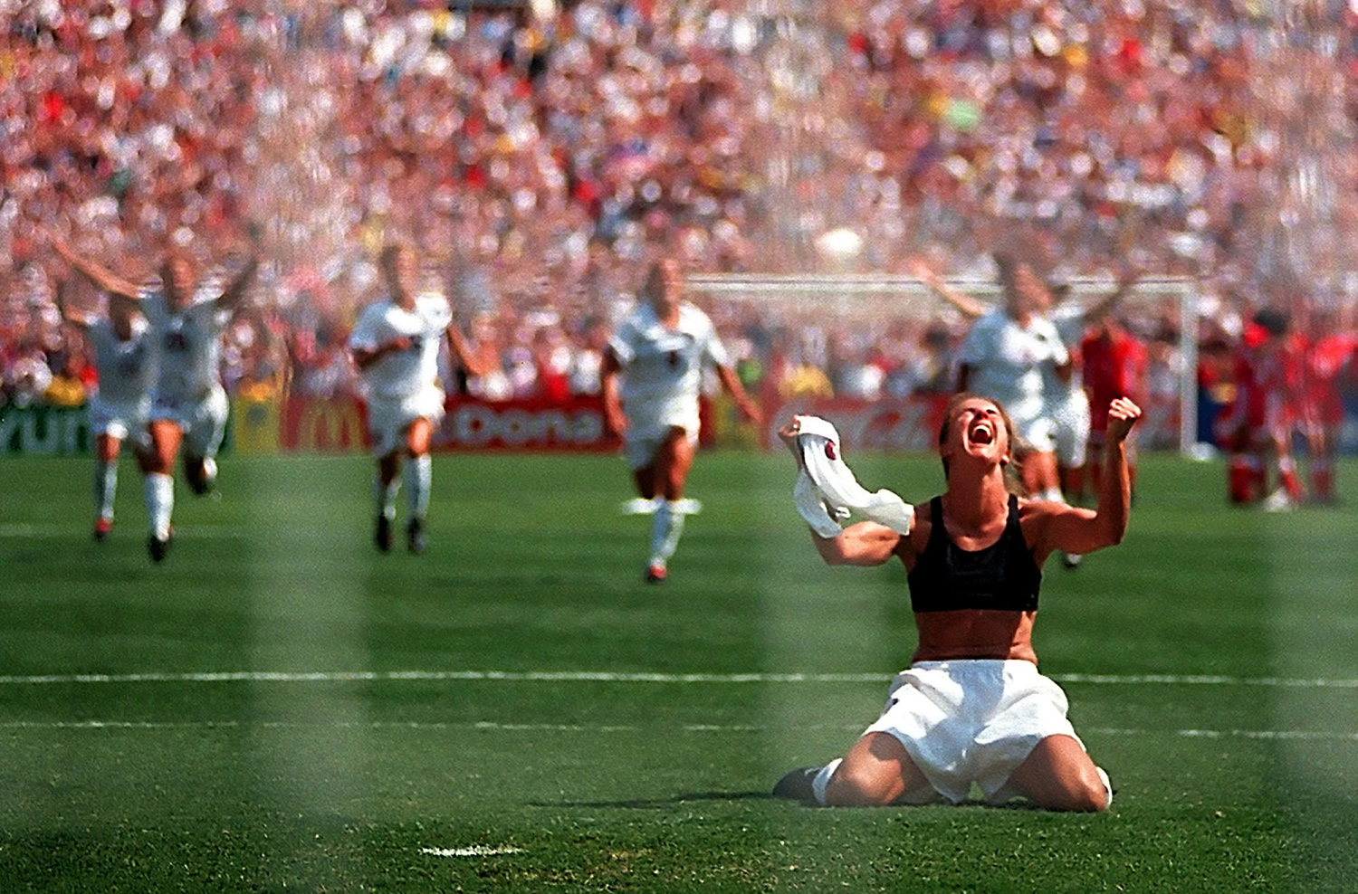 Brandi Chastain cheers on a field while baring her sports bra. Behind, her teammates run to meet her.