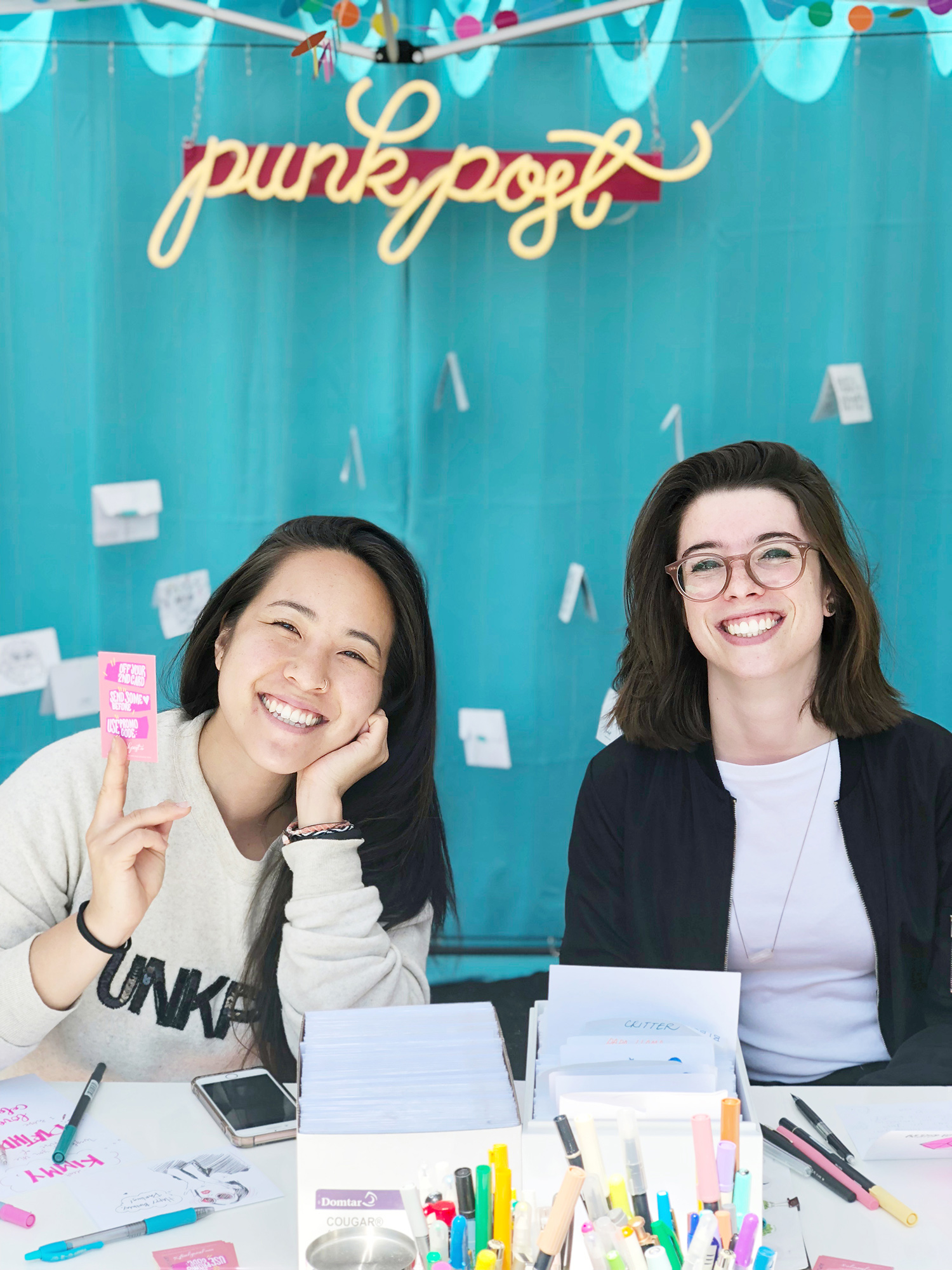 Two Punkpost artists smiling in an outdoor market booth with colorful markers and card making supplies. 
