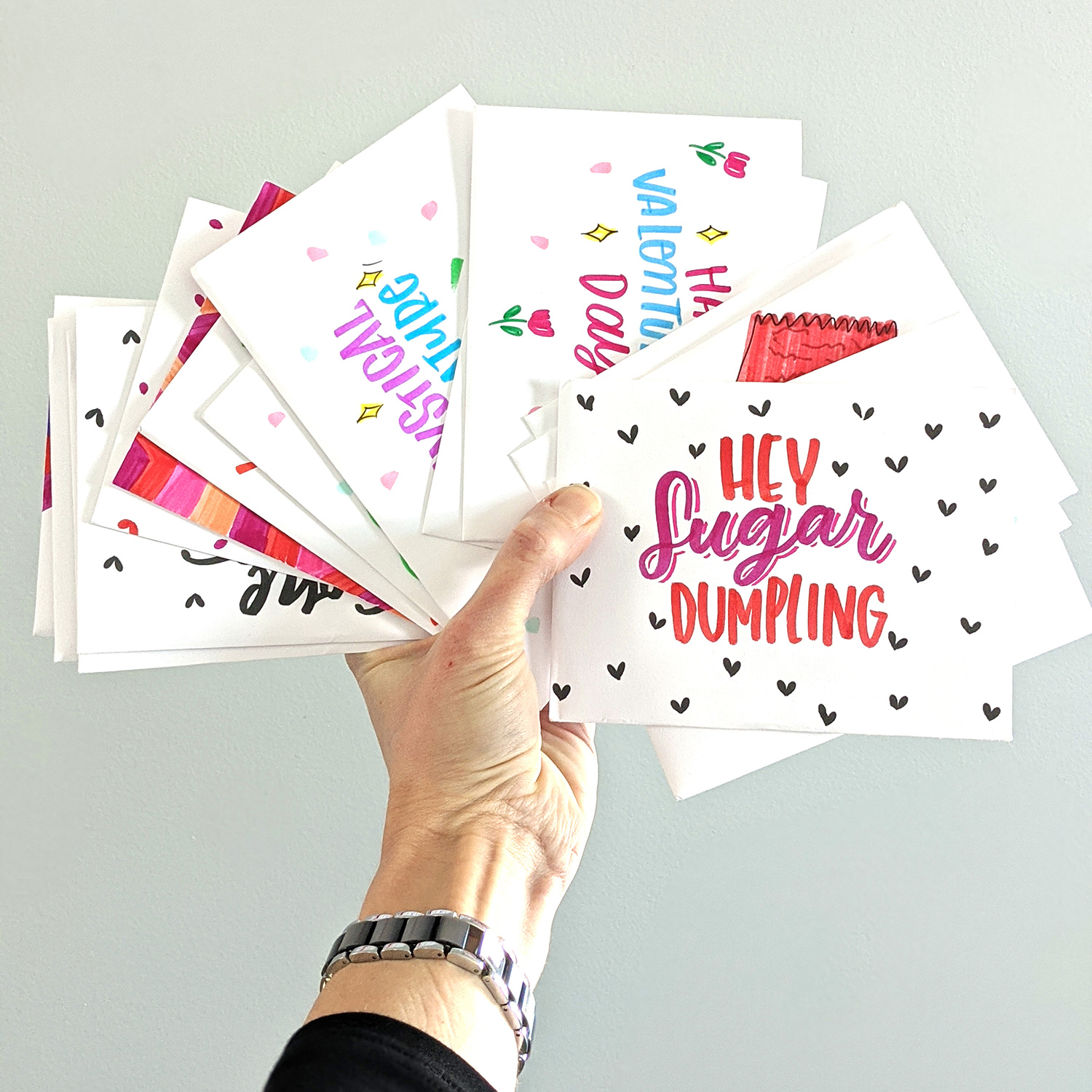 A hand holding many colorful Valentine’s Day cards fanned out. The top card reads “Hey sugar dumpling,” and is decorated with hearts. 