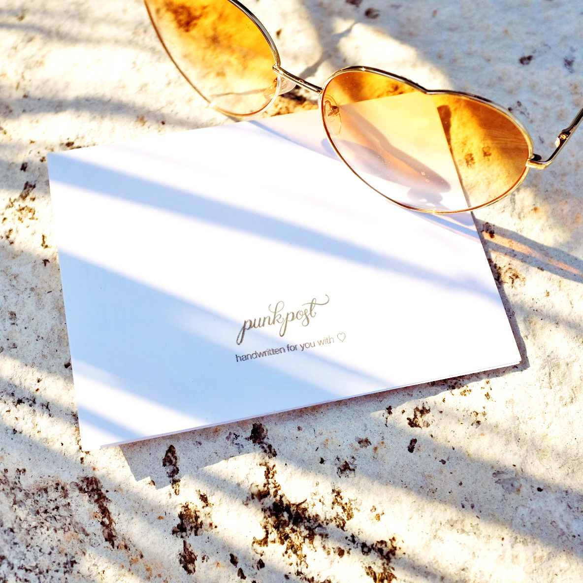 A face down greeting card with orange, heart-shaped sunglasses. Small lettering on the card reads “punkpost” in scripted font and “handwritten for you with” and a heart. 