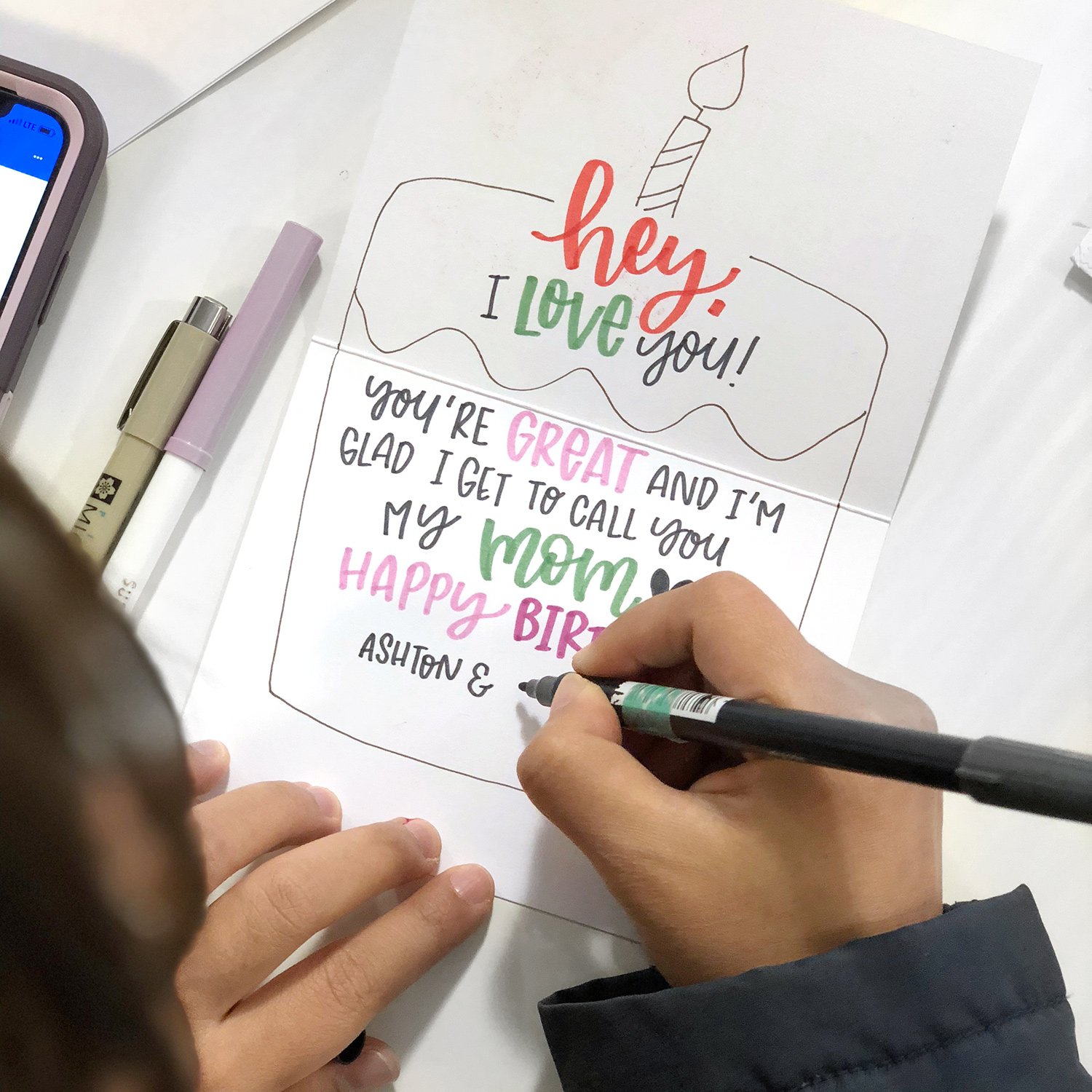 An artist writes a mother’s day message inside a card, using bright, fun lettering and a drawing of a cake with a candle.
