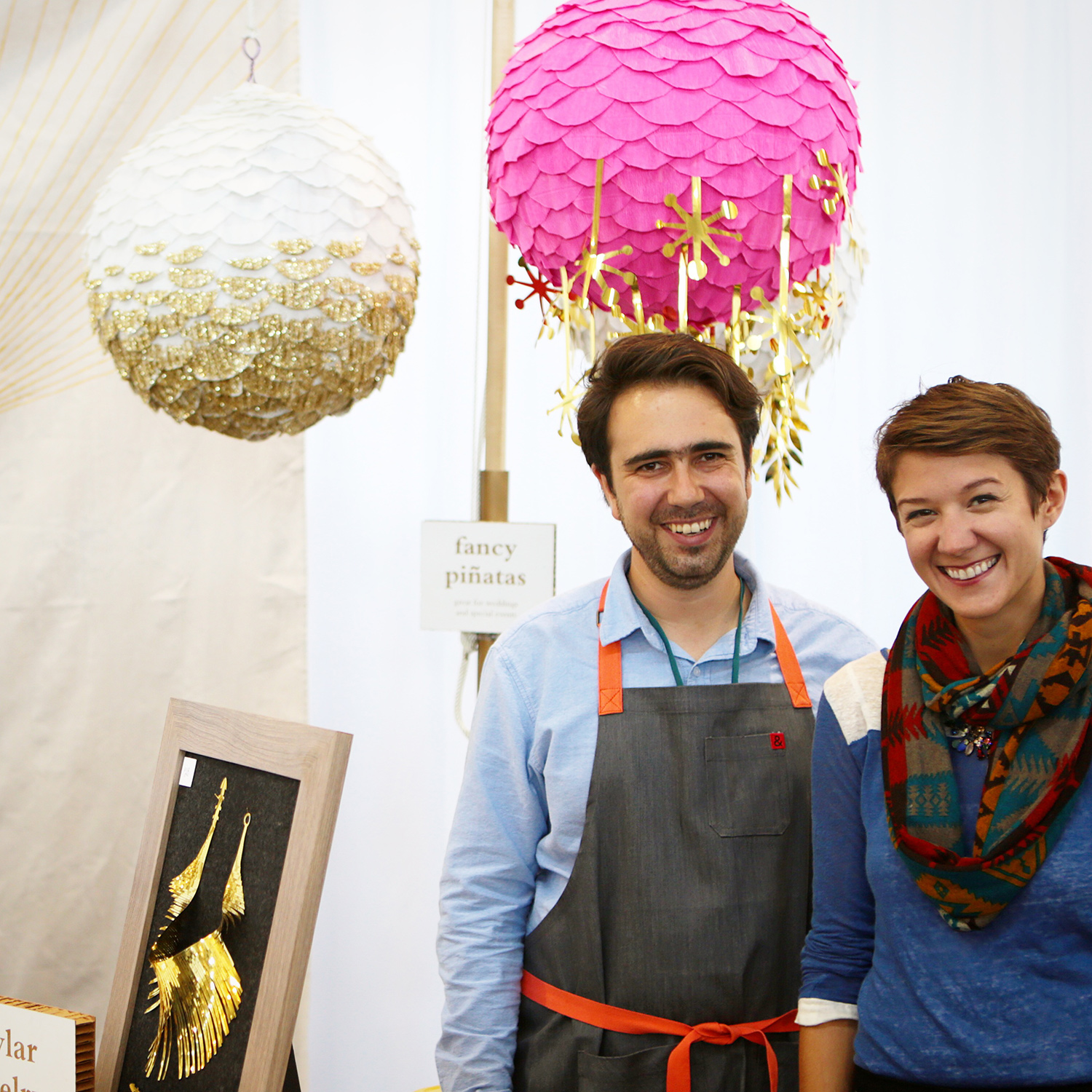 Lex and Santiago smile in an outdoor booth with piñatas and party decorations for sale. 