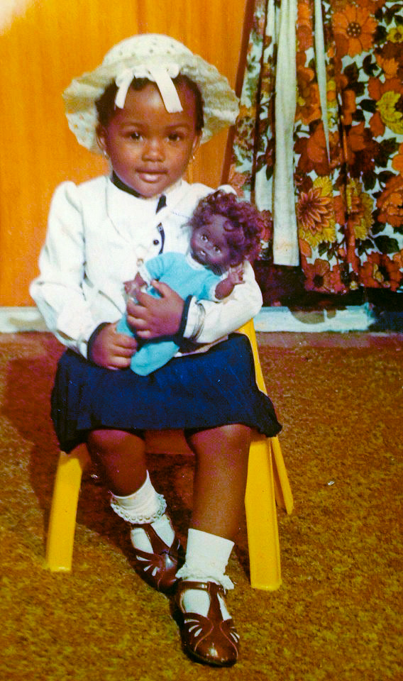 Arlyne, pictured as a small child, holds a doll and sits on a yellow stool.