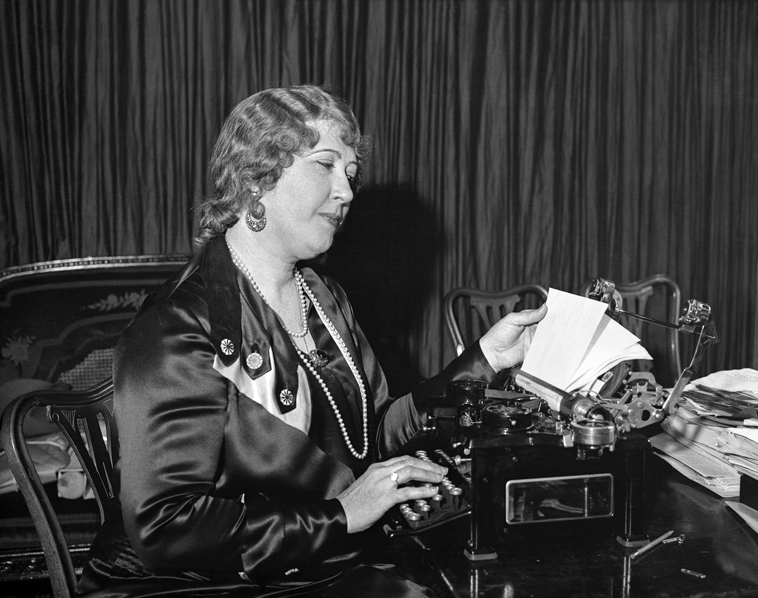 A middle-aged woman in a silk dress sitting in a chair while examining a typewriter