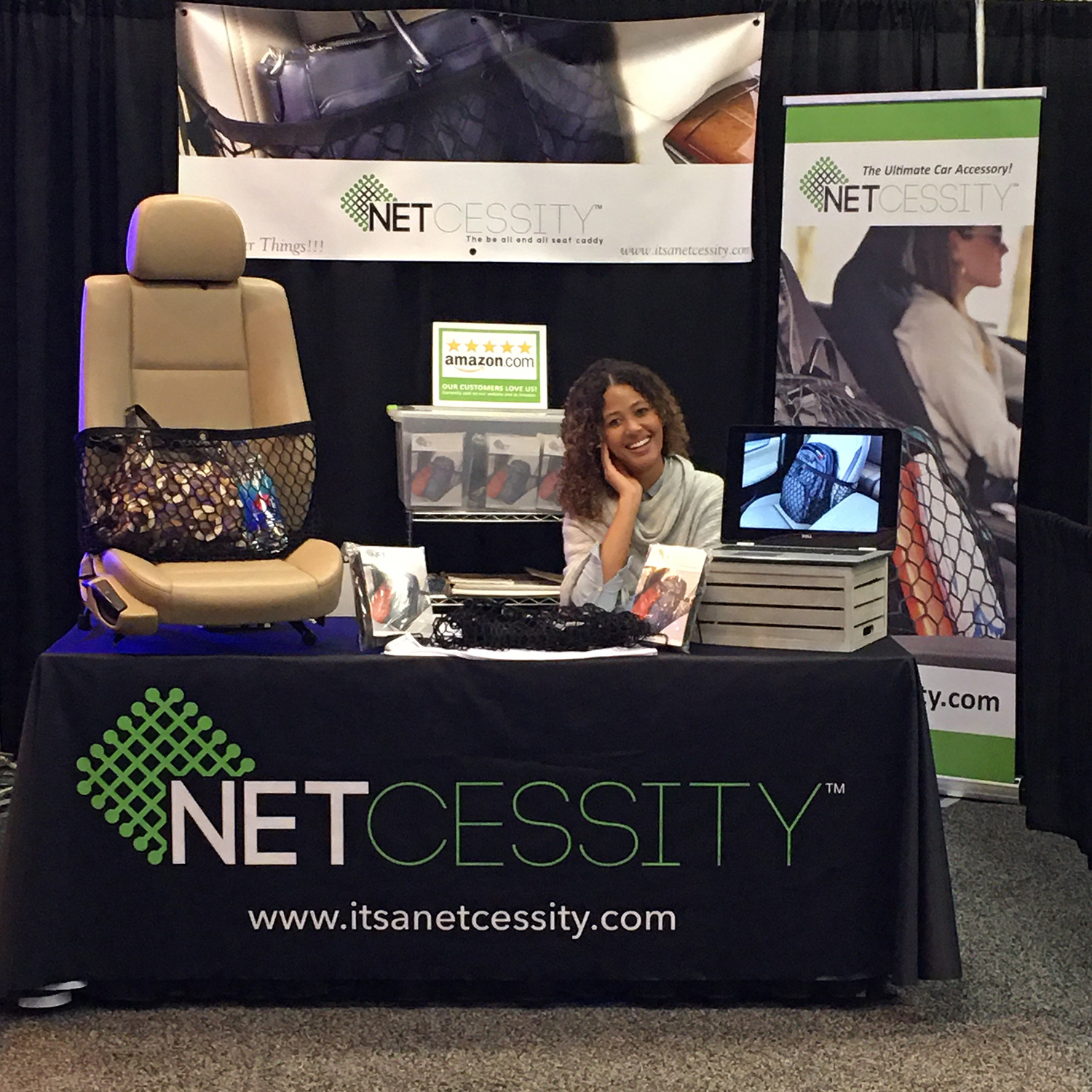 A smiling woman sits at a booth with a black tablecloth that says “Netcessity” and has a carseat with the Netcessity product on it and items inside the net. Banners surround the booth.