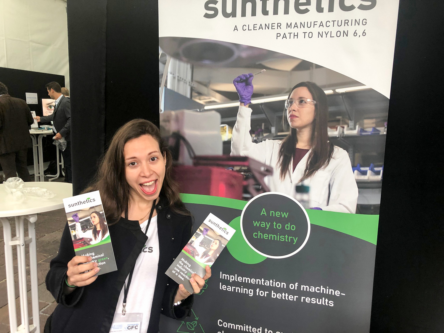 Daniela Blanco excitedly holds up company brochures while a promotional banner shows Daniela in the lab and reads: “A cleaner manufacturing path to Nylon 6,6”, “A new way to do chemistry”, and “Implementation of machine-learning for better results.”