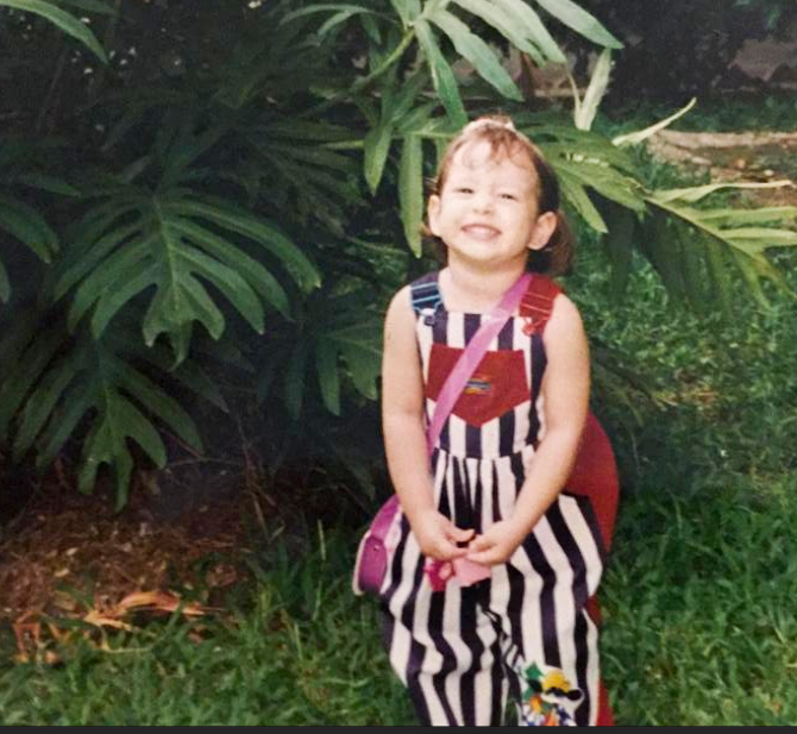 Daniela Blanco, a 1990s pre-school aged child, prepares to go to school wearing an oversized backpack and colorful striped overalls.