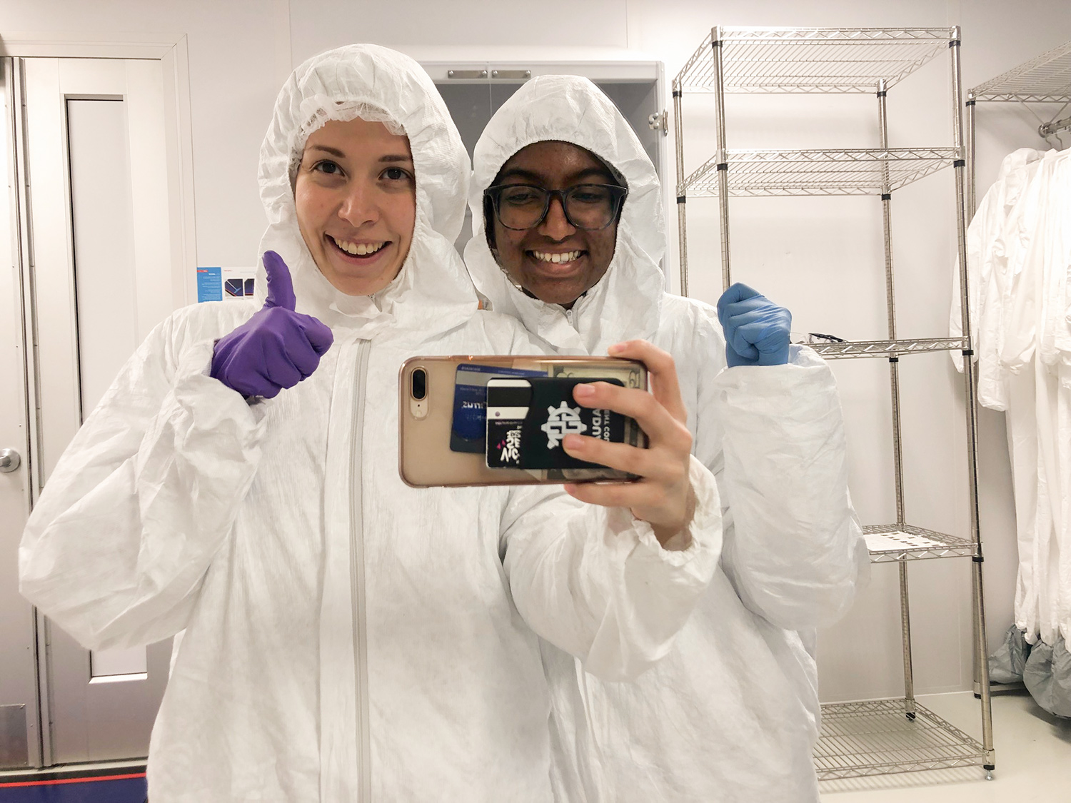Wear white ‘clean room’ jumpsuits that cover everything but their faces, Daniela Blanco and a dark-skinned woman stand in a lab smiling at the camera. Daniela gives a “thumbs up” with one hand while taking a cellphone self-portrait with each other.