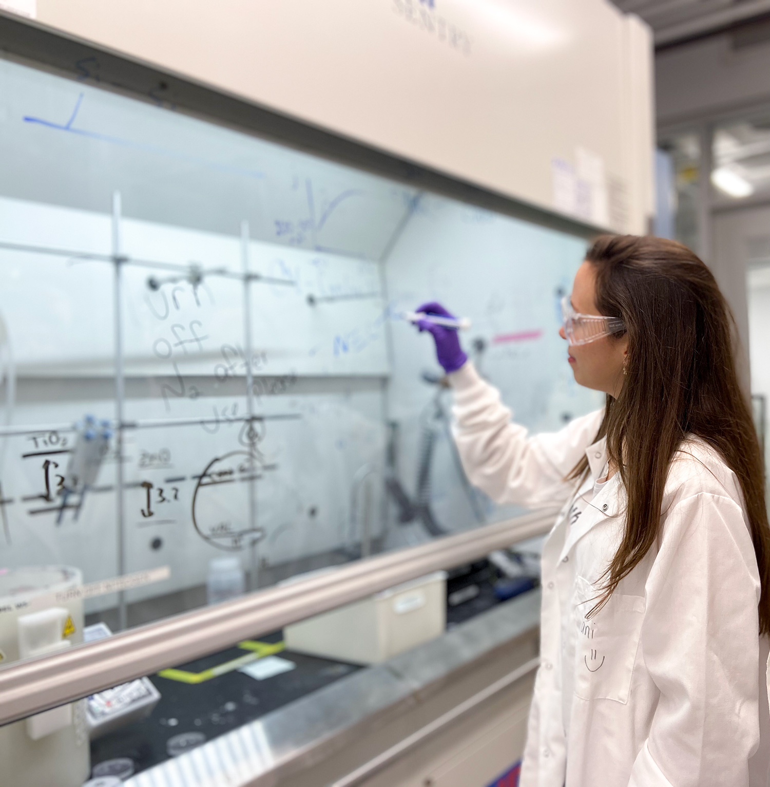 Chemical Engineer Daniela Blanco intently writes out chemical equations on a glass window inside a chemistry lab. She wears traditional lab attire – white lab coat, safety goggles, and plastic gloves.