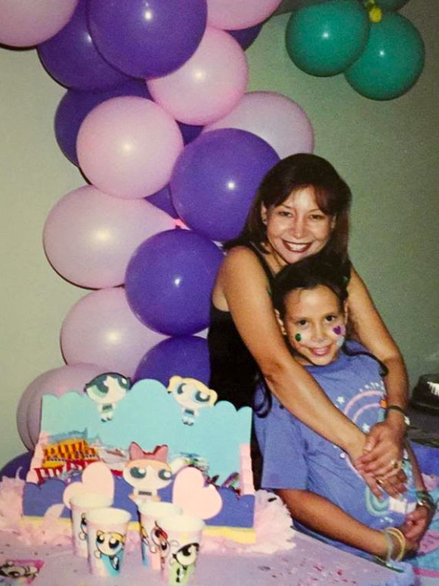 En una instantánea familiar de la década de 1990, la madre de Daniela Blanco envuelve a su hija Daniela, en edad de escuela primaria, en un abrazo. Se encuentran junto a un pastel y un arco de globos.