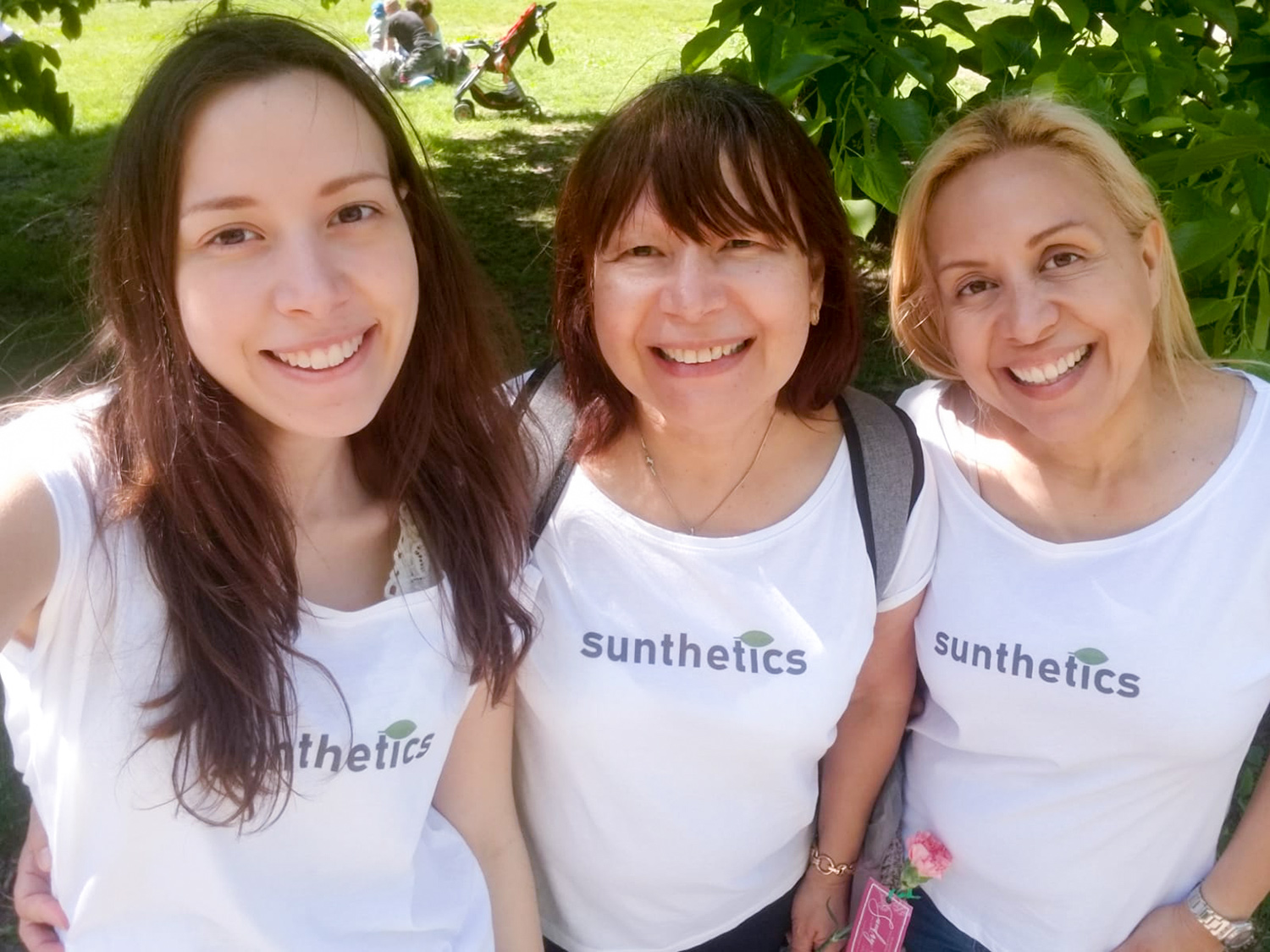 Daniela Blanco and two women take a selfie, all wearing Sunthetics company t-shirts. The logo on the t-shirt is the word “Sunthetics” in modern brown-gray font, with a green stylized leaf over the “i” of “Sunthetics.”