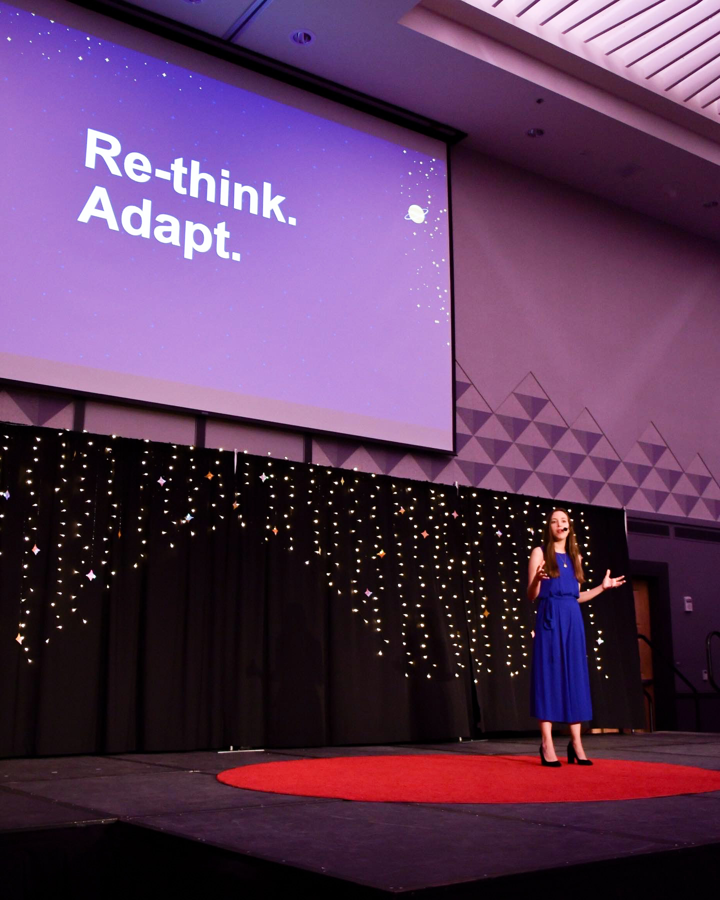 Daniela stands on a darkened stage within a red circle of carpet while giving a TED Talk. Above her head is a large presentation screen displaying bold white letters reading: “Re-think. Adapt.”