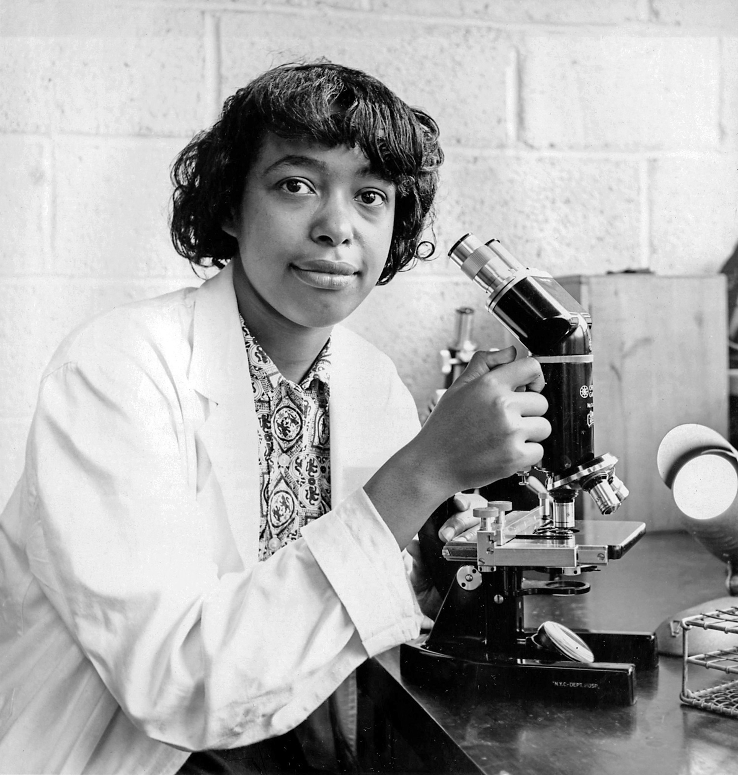 A teenager, the future Dr. Patricia Bath adjusts a microscope and looks into the camera. 