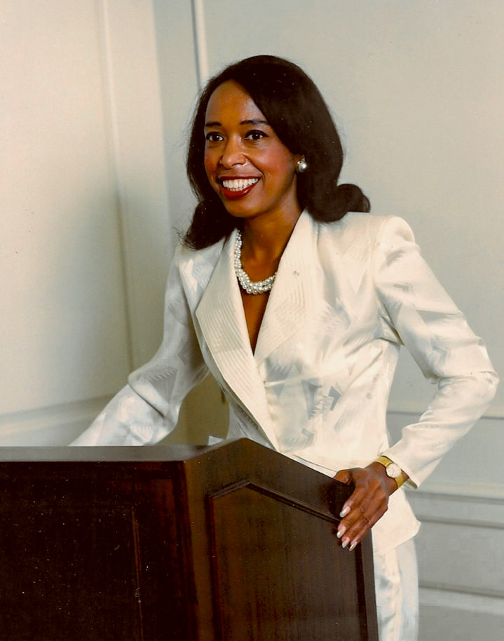 Dr. Patricia Bath stands smiling at a lectern and wears a fitted white satin suit. 