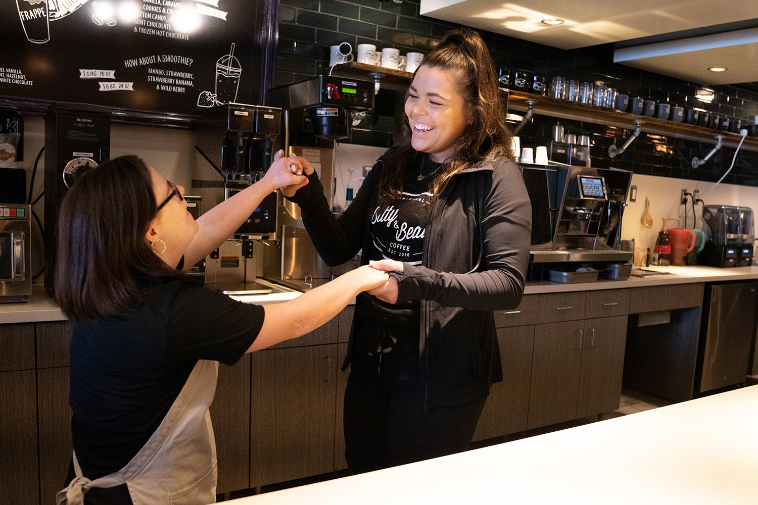 Two female employees smiling and dancing behind the serving counter. The coffee equipment work area behind them is clean and tidy.