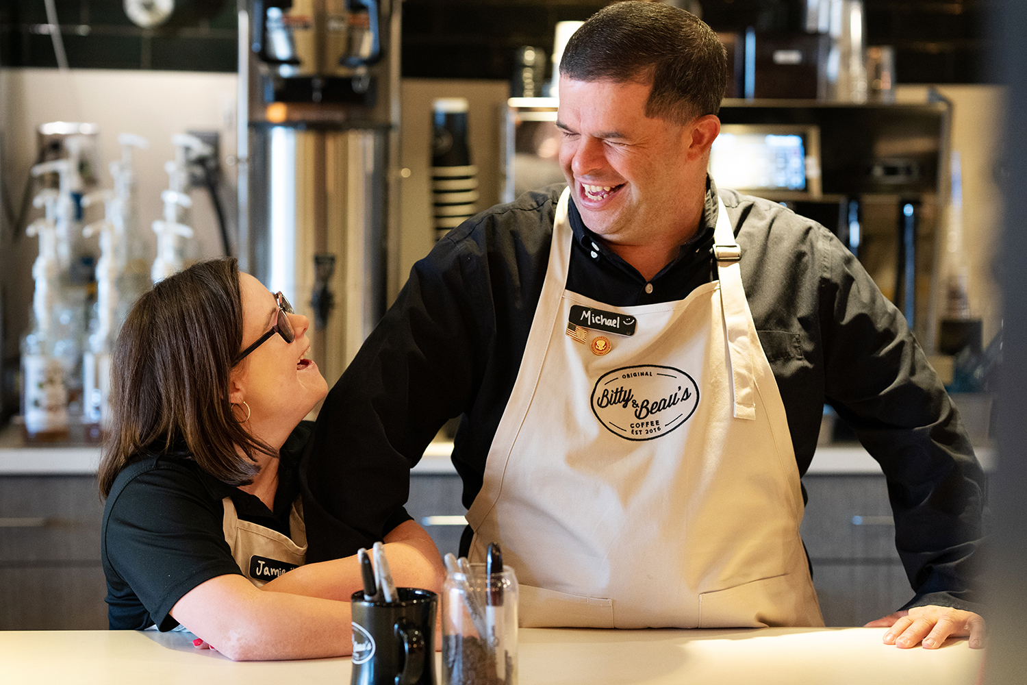 Two employees stand side by side at a counter wearing beige aprons with the Bitty and Beau's logo. They smile at each other and link arms. Coffee machines are in the background. 