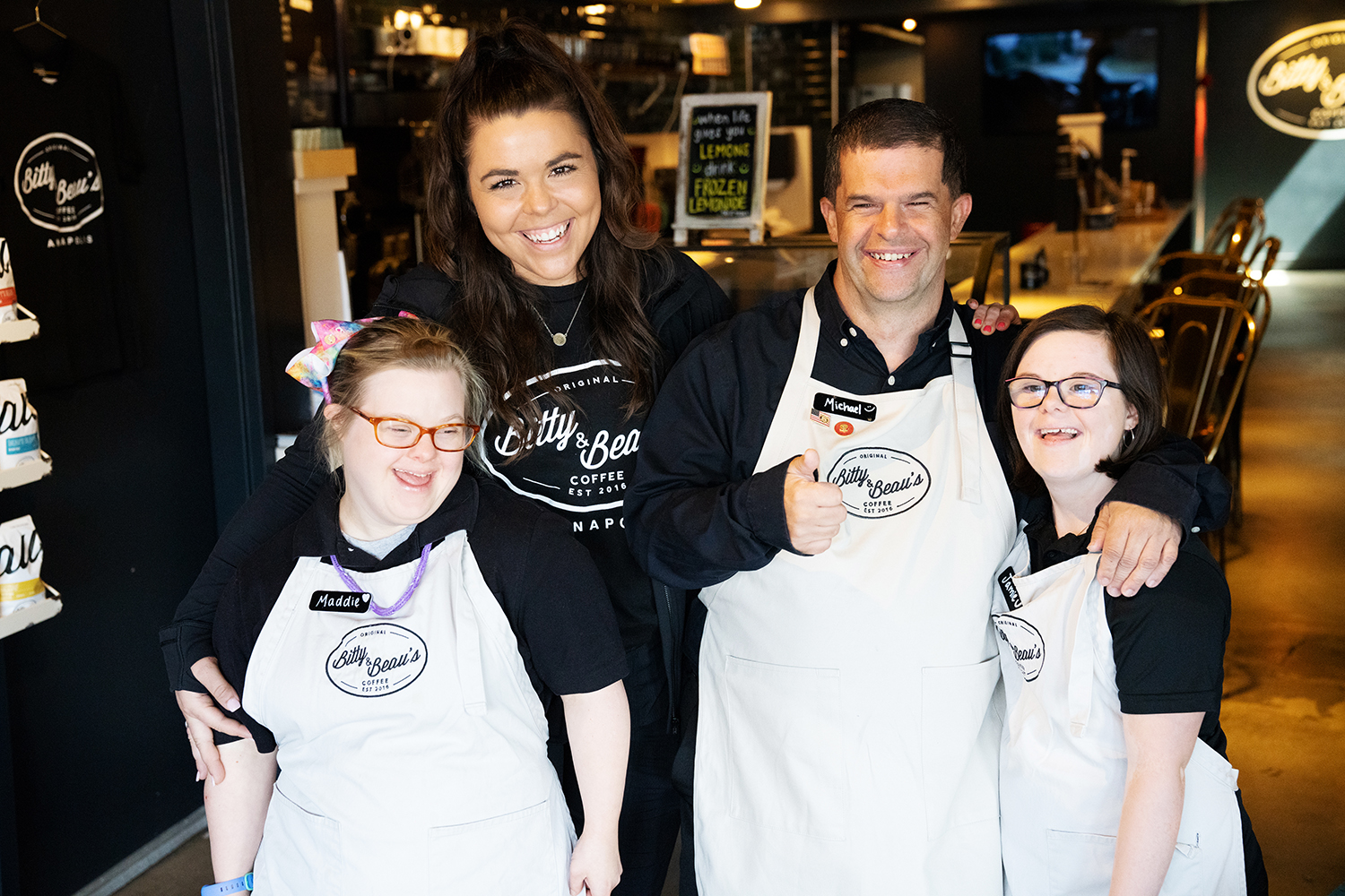 Four employees smile at the camera with arms around each other inside the shop. Male employee gives a thumbs up. All wear black shirts. Three wear beige aprons with a black logo.