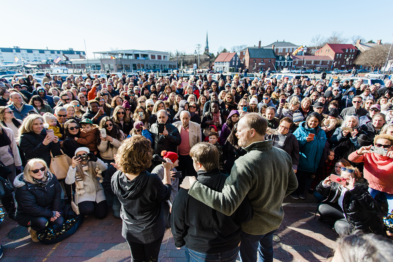 On a sunny day, Amy, Beau, and Ben Wright face a crowd on a street. Amy holds a microphone and Beau and Ben turn slightly toward her. The crowd is engaged, some holding up smartphones with cameras facing the Wrights. Many people wear warm clothing.