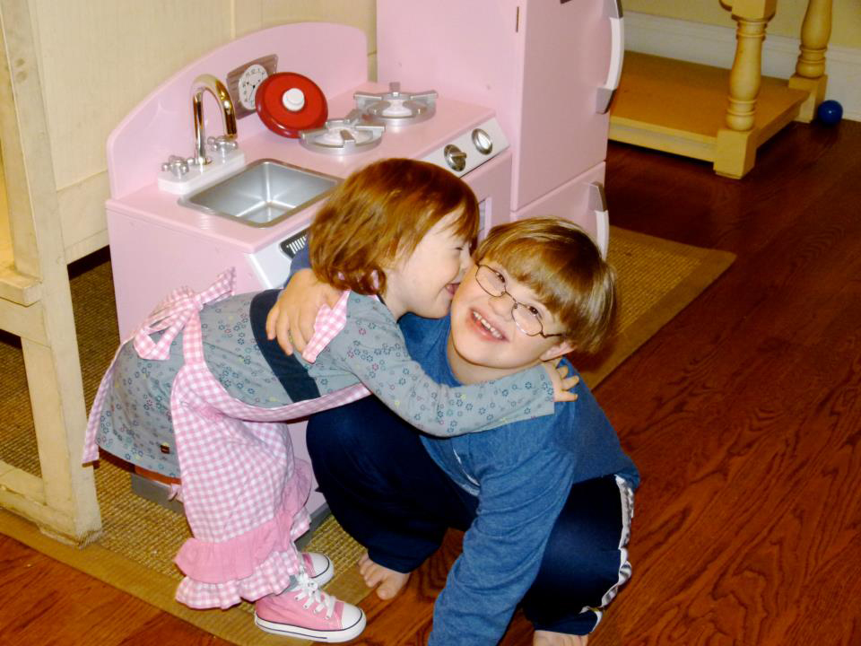 Boy, about eight years old, squats on the floor and smiles with one arm around a girl, about three years old. She leans over with her arms around his neck, smiling and kissing him on the cheek. A pink toy kitchen set is behind them. 