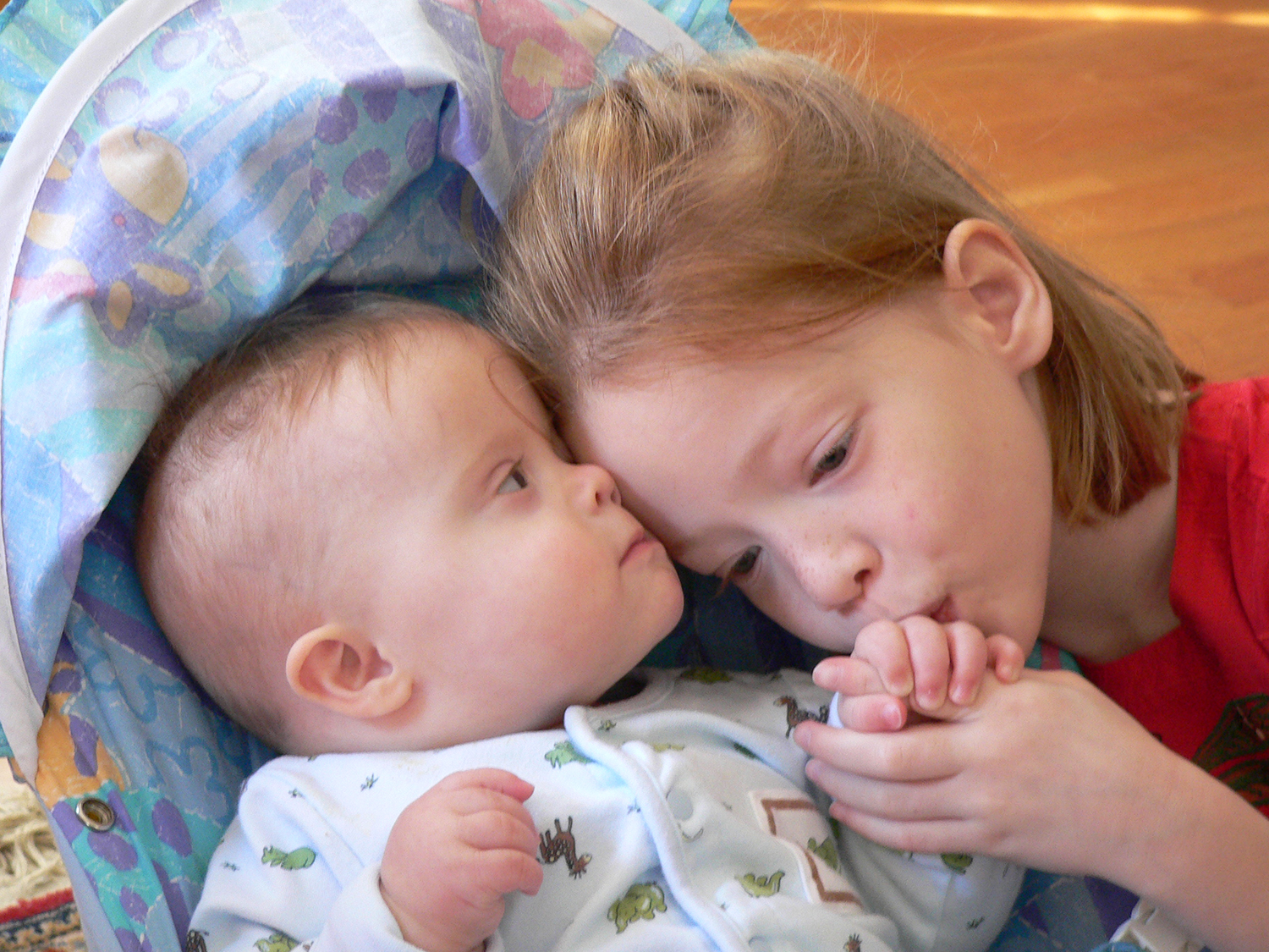 An infant faces a young girl, about six years old. She snuggles her head to his and kisses his hand while he grips her index finger. 