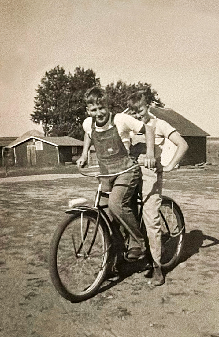 Black and white photo from the 1950s with two teenage boys in overalls riding a bike together: Al Kurtenbach standing in front holding the handlebars and Denis Kurtenbach in back sitting on the seat. Photo courtesy of Al Kurtenbach.