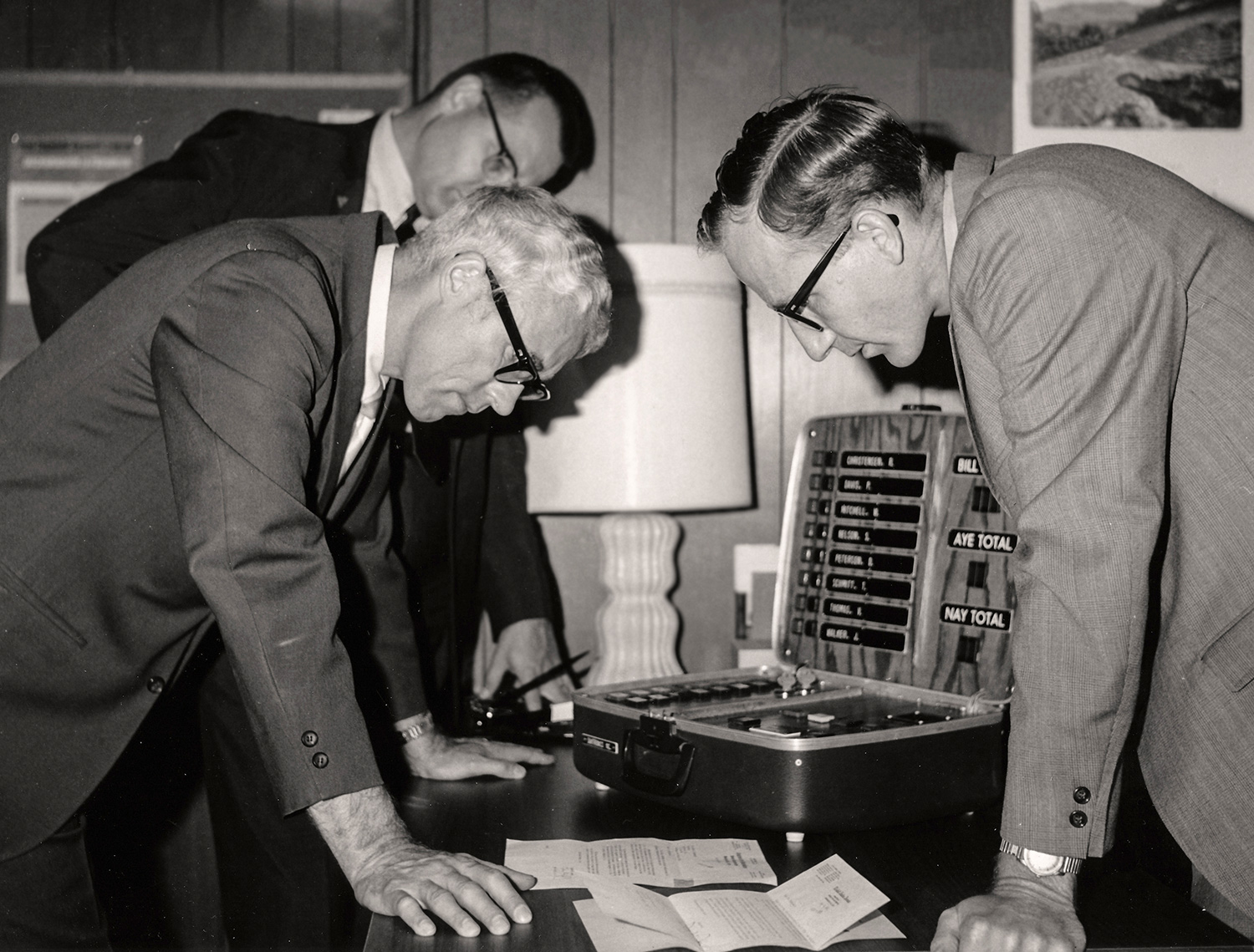 Black and white photo from the 1970s with three men in suits and ties in an office look at paperwork and an electronic voting machine on a table in an office. Left, Governor Frank Farrar, unknown man behind governor, Al Kurtenbach on the right. Photo courtesy of Daktronics.