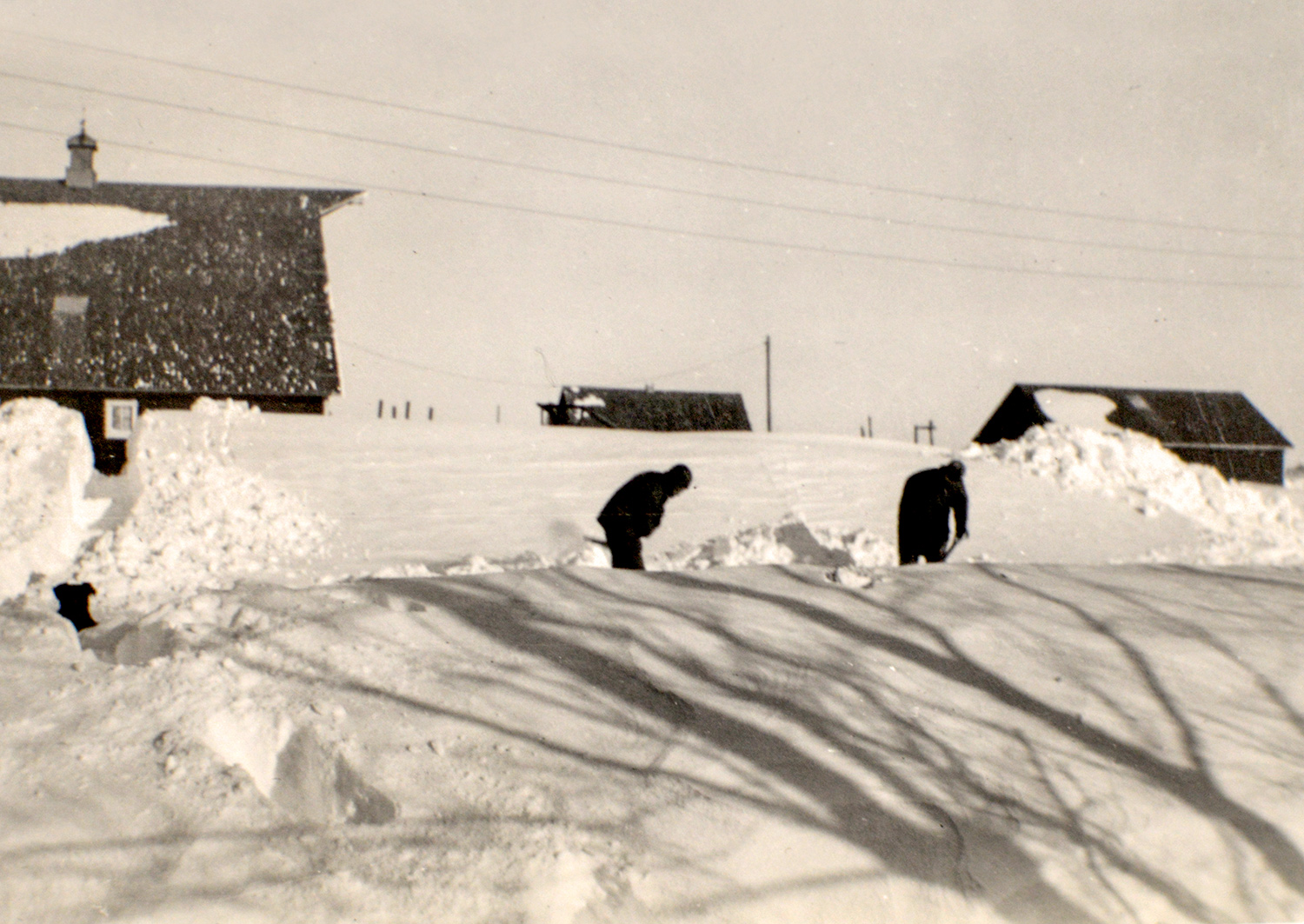Black and white photo from the 1950s with teenager Duane Sander and a friend shoveling a path through knee-deep snow with farm buildings in the background surrounded by tall snowdrifts. Photo courtesy of Duane Sander.