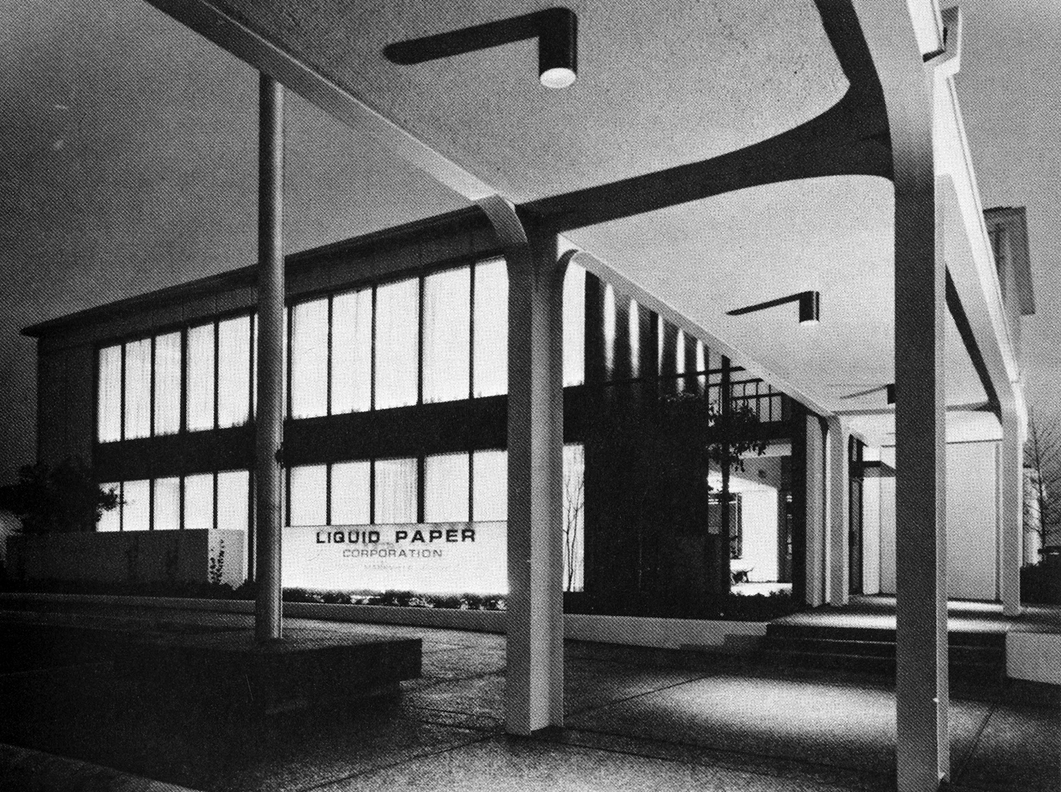 Black and white photograph showing front entrance view of Liquid Paper headquarters and manufacturing plant. The multistory building is made of concrete with a glass façade. 