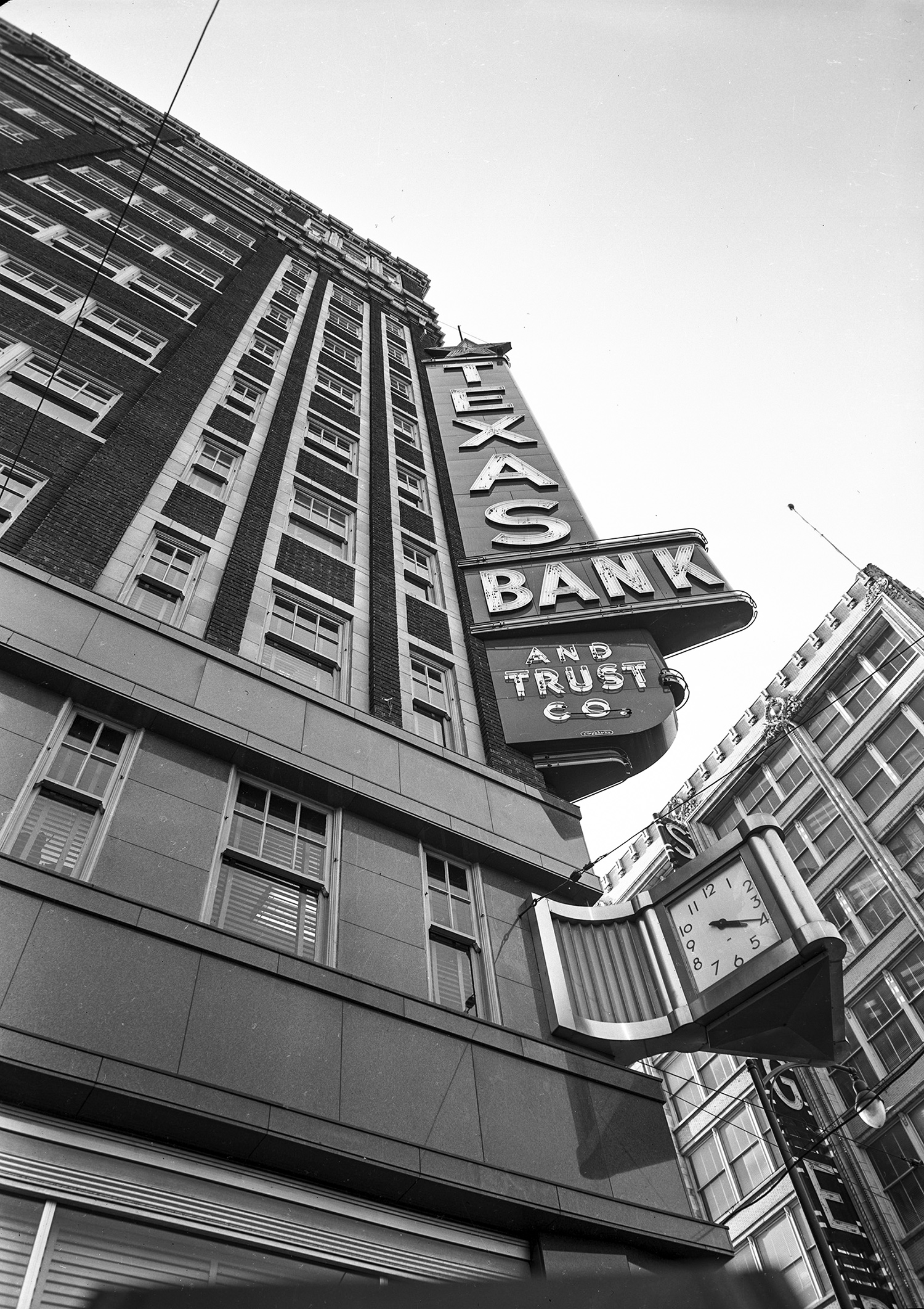 A black and white photograph of a multistory bank building in downtown Dallas, Texas, taken in the 1950s. The photograph is a vertical shot showing the brick and stone façade and multitude of windows. 
